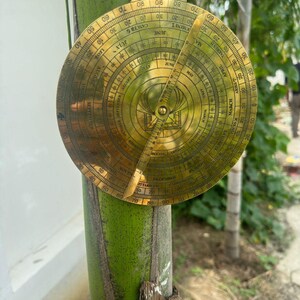 May include: A vintage brass astrolabe, a historical astronomical instrument, is displayed. The circular device features intricate engravings, including degree markings, zodiac signs, and months. A pointer extends from the center, and a hanging loop is at the top.