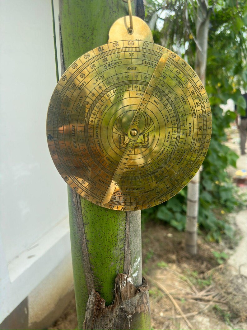 May include: A vintage brass astrolabe, a historical astronomical instrument, is displayed. The circular device features intricate engravings, including degree markings, zodiac signs, and months. A pointer extends from the center, and a hanging loop is at the top.