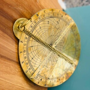 May include: A vintage brass astrolabe with intricate markings and a chain. The circular instrument features a rotating dial with degree markings and a central pointer. The astrolabe is a historical astronomical instrument.