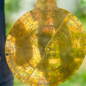 Puede incluir: Un astrolabio de latón antiguo, un instrumento astronómico histórico, con intrincadas marcas grabadas y una regla giratoria. El diseño circular incluye signos del zodiaco, meses y escalas de grados. El instrumento está sostenido por una mano.
