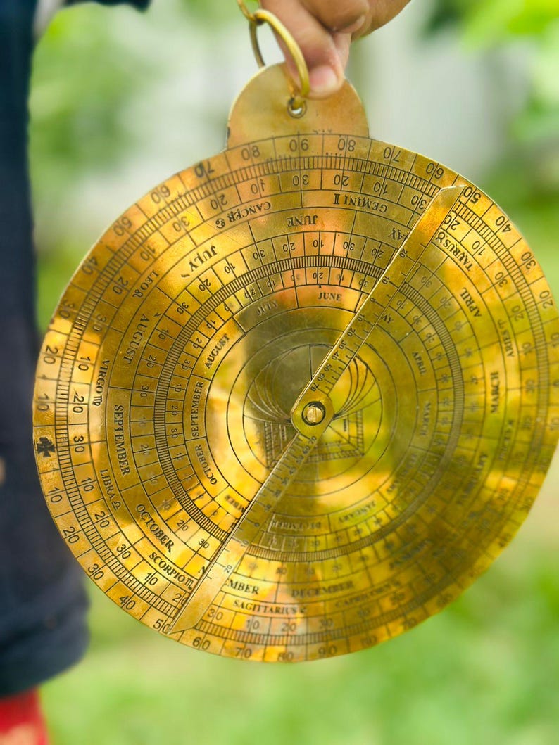 May include: A vintage brass astrolabe, a historical astronomical instrument, featuring intricate engraved markings and a rotating rule. The circular design includes zodiac signs, months, and degree scales. The instrument is held by a hand.