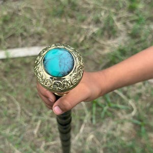 Op de afbeelding: Decoratieve wandelstok met een goudkleurige handgreep met een sierlijk ontwerp en een turkooiskleurige steen. De schacht van de stok is zwart met een spiraalpatroon, wat wijst op een vintage of antieke stijl.