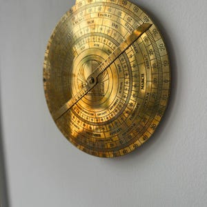 May include: A vintage brass astrolabe, a historical astronomical instrument, hangs against a white wall. The circular device features engraved markings, including months, zodiac signs, and degree scales. A rotating arm points to the current date.