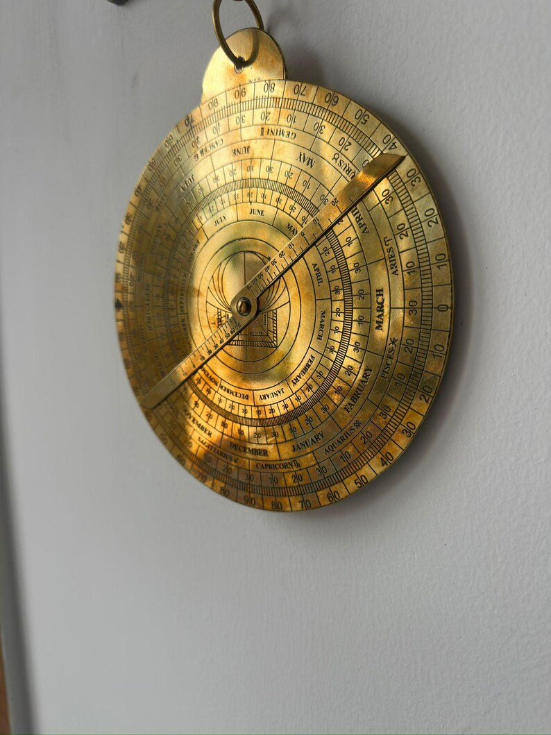 May include: A vintage brass astrolabe, a historical astronomical instrument, hangs against a white wall. The circular device features engraved markings, including months, zodiac signs, and degree scales. A rotating arm points to the current date.