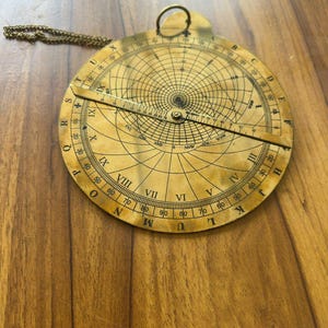 May include: A vintage brass astrolabe with intricate markings and a rotating arm. The circular instrument features Roman numerals, degree markings, and a chain for hanging. The astrolabe is a navigational tool.