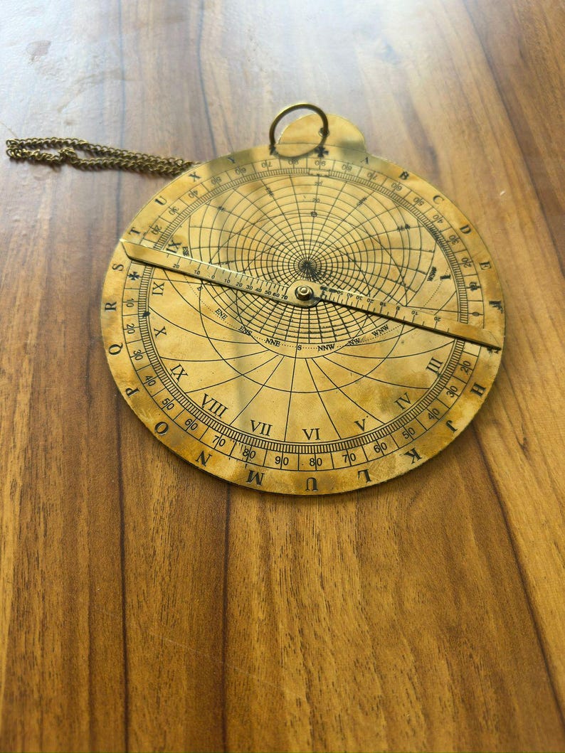 May include: A vintage brass astrolabe with intricate markings and a rotating arm. The circular instrument features Roman numerals, degree markings, and a chain for hanging. The astrolabe is a navigational tool.