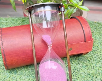 May include: An hourglass with a clear glass chamber filled with pink sand. The hourglass is held in a bronze-colored frame. A red leather case rests behind the hourglass on a bed of green grass.
