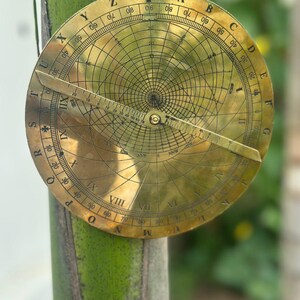 May include: A vintage brass astrolabe, a historical astronomical instrument, featuring intricate engraved markings and a rotating pointer. The circular design includes degree scales and celestial coordinates, with a small loop at the top for hanging.