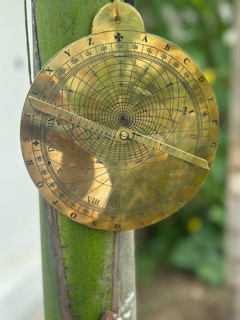 May include: A vintage brass astrolabe, a historical astronomical instrument, featuring intricate engraved markings and a rotating pointer. The circular design includes degree scales and celestial coordinates, with a small loop at the top for hanging.