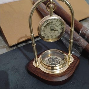 May include: A brass and wood desk clock with a compass and a clock face that reads "Victoria London 1876". The clock has a gold-coloured arch and is set on a dark wood base. The clock is a decorative item.