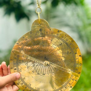 May include: A vintage brass astrolabe, a historical astronomical instrument, is suspended by a gold chain. The circular device features intricate markings, including degree scales, Roman numerals, and a central pointer. The background is a blurred green, suggesting an outdoor setting.