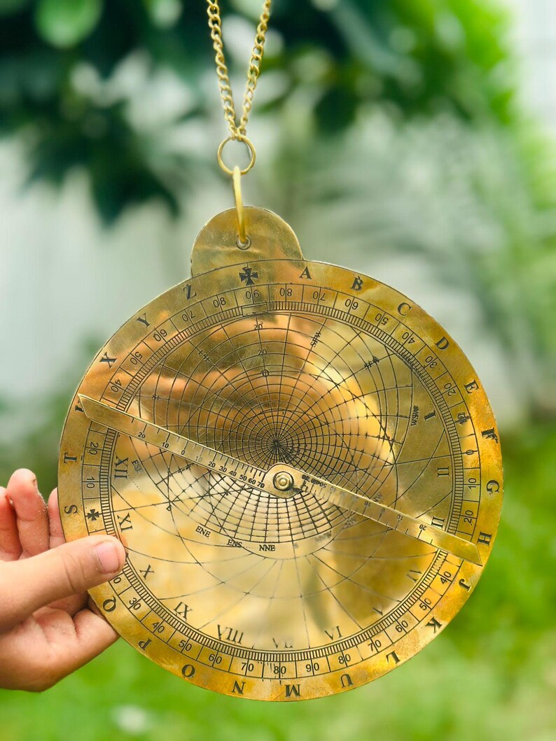 May include: A vintage brass astrolabe, a historical astronomical instrument, is suspended by a gold chain. The circular device features intricate markings, including degree scales, Roman numerals, and a central pointer. The background is a blurred green, suggesting an outdoor setting.
