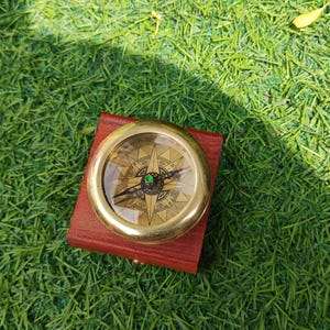 Peut inclure: Une boussole en laiton vintage avec un cadran détaillé, posée sur un petit étui en cuir rouge. La boussole est placée sur un fond d'herbe verte vibrante, suggérant une utilisation en extérieur ou un thème nautique. La boussole a un couvercle en verre transparent.