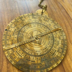May include: A vintage brass astrolabe with engraved markings, including months, zodiac signs, and degree measurements. The circular instrument features a rotating arm and a chain for hanging. The astrolabe is a historical astronomical device.