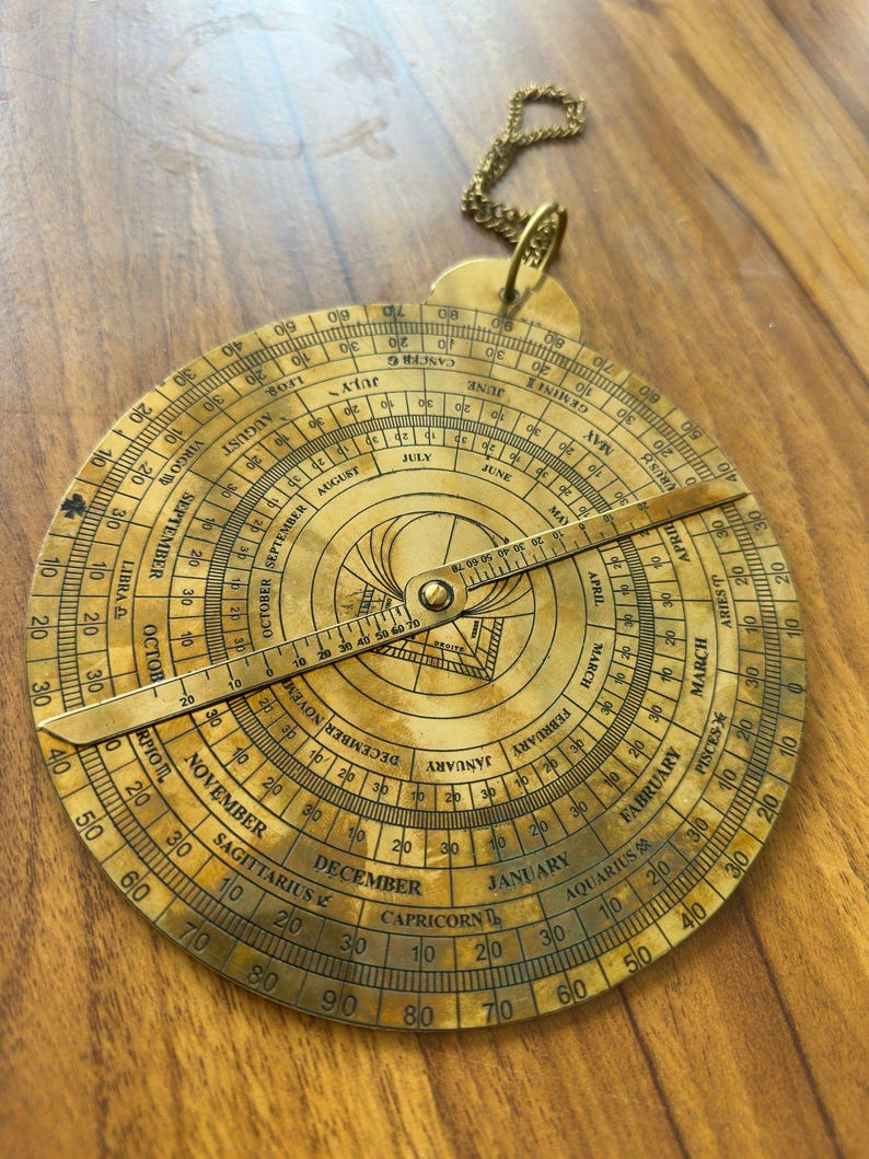 May include: A vintage brass astrolabe with engraved markings, including months, zodiac signs, and degree measurements. The circular instrument features a rotating arm and a chain for hanging. The astrolabe is a historical astronomical device.