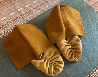 May include: A pair of mustard yellow leather moccasins featuring a soft, gathered toe design and a tall, folded-over cuff made of a darker, suede-textured brown leather. The footwear has a flexible construction with a smooth, matte finish.