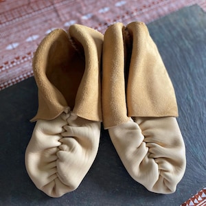 May include: A pair of soft-soled moccasins crafted from light beige leather. The footwear features a gathered, puckered toe design and a wide, folded-over cuff at the ankle opening, showcasing a smooth texture on the vamp and a suede finish on the cuff.
