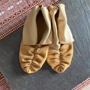 May include: A pair of soft, tan-colored leather moccasins featuring a gathered, puckered toe design and a lighter, smooth cream-colored leather cuff that extends upward from the opening of each shoe, showcasing a natural, unlined texture.