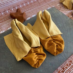 May include: A pair of soft-soled moccasins crafted from mustard yellow suede leather. The footwear features a gathered toe design and a wide, folded-over cuff in a lighter shade of pale yellow, showcasing a textured, matte finish throughout.