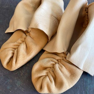 May include: A pair of light beige moccasins featuring a soft, gathered toe construction and a high, folded cuff. The footwear is crafted from a textured, natural-toned material with visible stitching details along the center of the toe box.