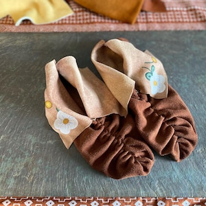 May include: A pair of brown baby booties with a gathered design. The booties have a light tan cuff with embroidered white flowers and green leaves. A small gold button is on the side of each bootie.