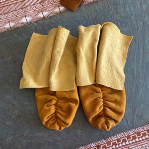 May include: A pair of soft leather moccasins featuring a two-tone design with golden-yellow suede vamps and lighter tan leather cuffs. The footwear has a gathered, ruched toe construction and a flexible, unlined structure with visible seams.