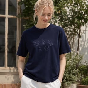 May include: A navy blue short-sleeved t-shirt featuring a subtle, tonal graphic print of various seashells and starfish arranged in a cluster on the chest. The garment has a classic crew neckline and a straight hem, providing a simple, clean look.