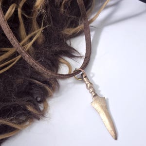 May include: A bronze spearhead pendant on a brown leather cord. The pendant is attached to a small gold-coloured ring. The background includes brown and blonde fur.