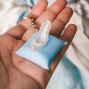 May include: A miniature translucent white high-heeled shoe keychain featuring a small bow detail on the toe, securely attached to a silver-toned metal keyring and resting on a small, square, light blue pillow with a smooth, matte finish.