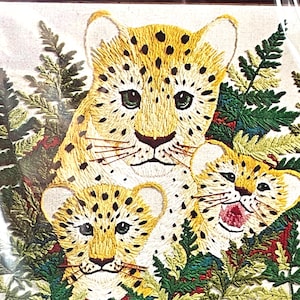 May include: An embroidery art piece on light fabric featuring a large leopard and two smaller leopard cubs nestled among detailed green fern leaves. The leopards have yellow fur with black spots, dark eyes, and whiskers, while one cub has an open mouth.