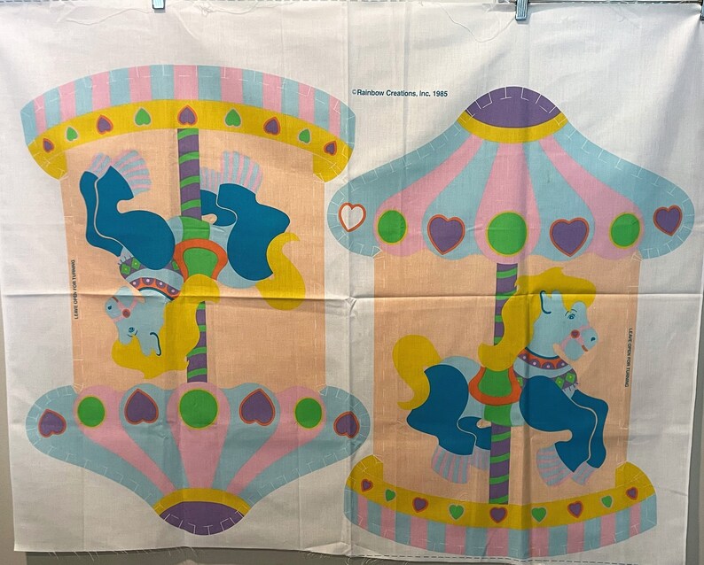 May include: A vintage fabric panel featuring two carousel horses with colorful details. The panel is divided into two sections, each with a carousel horse and a canopy. The canopies are decorated with hearts and stripes. The text "Rainbow Creations, Inc. 1983" is printed in the top right corner.