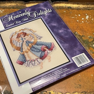 May include: A cross-stitch kit titled "Heavenly Delights" featuring a counted cross-stitch design of an angel. The kit includes 100% cotton 18 count Aida cloth, embroidery floss, metallic fibers, beads, needles, and instructions. The finished design measures 5 x 7 inches.