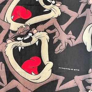 May include: Black fabric featuring a repeating pattern of the Tasmanian Devil cartoon character. The character is depicted with a wide open mouth, red tongue, and sharp teeth. The design includes the text "TM & © 1991 Warner Bros. Inc."