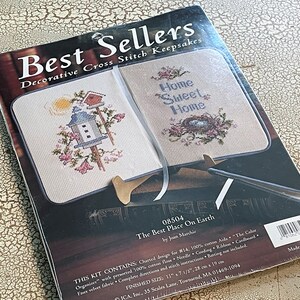 May include: A cross-stitch kit titled "Best Sellers" featuring two completed designs: a birdhouse with flowers and the text "Home Sweet Home." The kit includes floss, needle, and instructions. Finished size is 11" x 7 1/2".