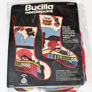 Könnte beinhalten: Ein Bucilla-Nadelset für ein Paar bemalter Weihnachtsstrümpfe. Die Strümpfe sind 24 cm lang und weisen farbenfrohe Designs mit einem Teddybären, einem Trommler und anderen Urlaubsbildern auf. Das Set enthält Wolle und Leinwand.