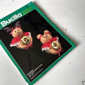 May include: A package of Bucilla Christmas Heirloom felt door knob covers. The covers are shaped like reindeer and teddy bears, with red and white accents, and gold-colored door knob covers. The package is green and black.