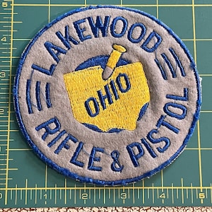 May include: A blue and gray embroidered patch with the text "Lakewood, Ohio Rifle & Pistol" and a yellow outline of the state of Ohio with a bullet inside.
