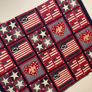 May include: A rectangular fabric wall hanging featuring a grid of squares with red, white, and blue patterns. The design includes American flag motifs, white stars on blue backgrounds, and red heart shapes containing eagle silhouettes and small buttons.