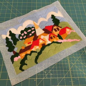 May include: Needlepoint landscape featuring a village with red-roofed houses, green hills, and a blue sky with white clouds. The needlepoint is on a white mesh canvas, set against a green cutting mat.