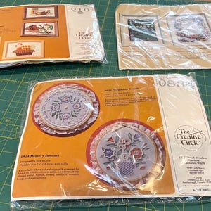 May include: Three vintage embroidery kits from The Creative Circle. The kits feature framed designs of floral arrangements, teapots, and baskets. The kits are packaged in clear plastic and are on a green surface.