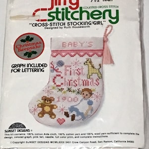 May include: A cross-stitch stocking kit featuring a baby's first Christmas theme. The stocking is pink and white with a giraffe, teddy bear, and a baby in a blue dress. The kit includes a graph for lettering, pink felt, needle, and full color print. "Christmas Keepsake" and "Baby's First Christmas" are printed on the packaging.  The finished size is 7 1/2 inches high.