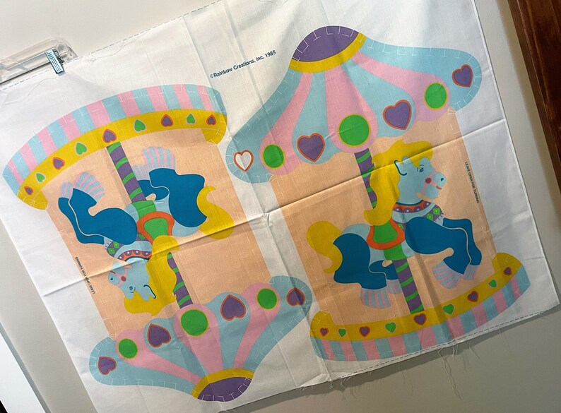 May include: A vintage fabric panel featuring a colorful carousel with two horses. The panel is printed with a light pink, blue, yellow, and green color scheme. The text "Rainbow Creations, Inc. 1985" is printed in the top right corner.