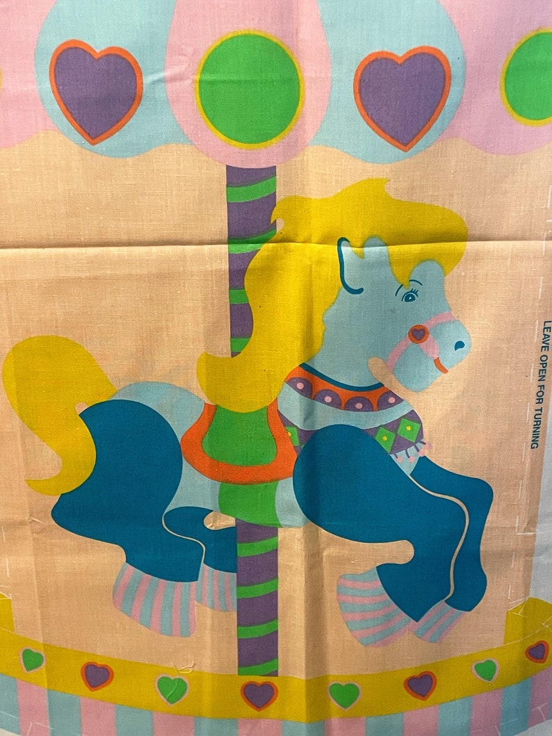 May include: A colorful fabric panel featuring a blue and yellow carousel horse with a striped pole and green, pink, and purple accents. The text "LEAVE OPEN FOR TURNING" is printed on the right side of the panel.