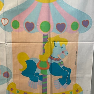 May include: A vintage fabric panel featuring a colorful carousel horse with a yellow mane and tail, blue body, and pink and white striped hooves. The carousel is decorated with hearts, circles, and stripes in pink, yellow, blue, green, and purple. The panel includes the text "Rainbow Creations, Inc. 1985" and "Leave open for turning".