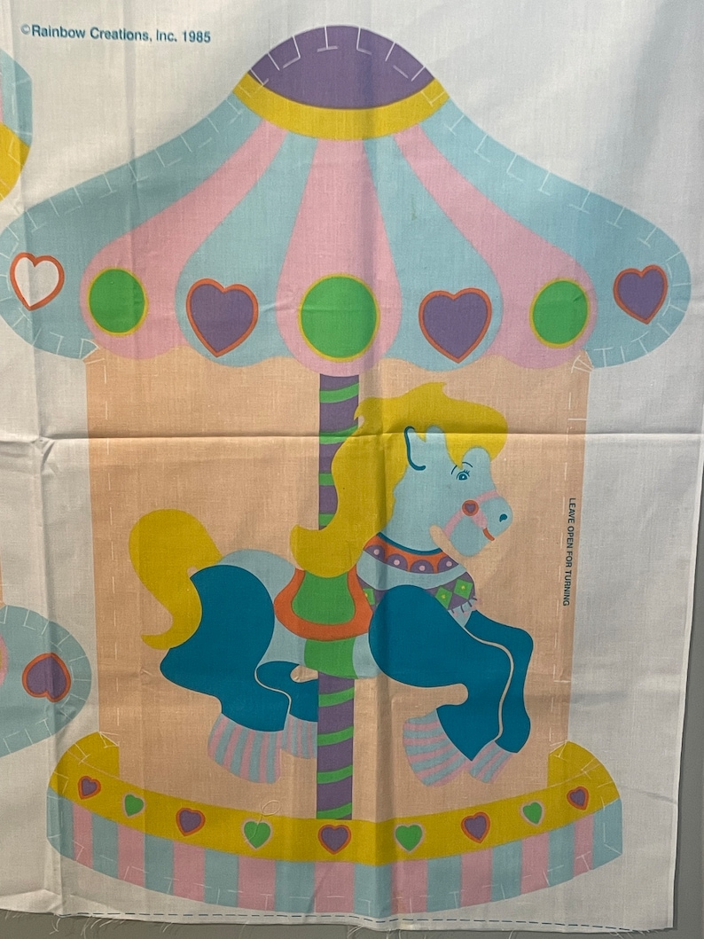 May include: A vintage fabric panel featuring a colorful carousel horse with a yellow mane and tail, blue body, and pink and white striped hooves. The carousel is decorated with hearts, circles, and stripes in pink, yellow, blue, green, and purple. The panel includes the text "Rainbow Creations, Inc. 1985" and "Leave open for turning".