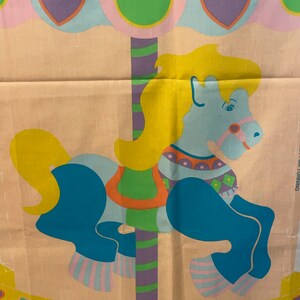 May include: A colorful fabric panel depicting a carousel horse with a blue body, yellow mane, and a striped pole. The horse is decorated with colorful geometric patterns. The background features pastel blue and pink clouds with hearts and green circles.