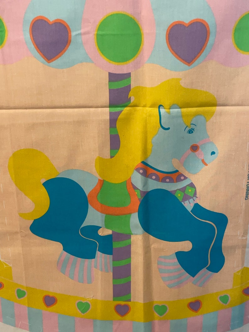 May include: A colorful fabric panel depicting a carousel horse with a blue body, yellow mane, and a striped pole. The horse is decorated with colorful geometric patterns. The background features pastel blue and pink clouds with hearts and green circles.