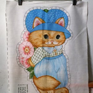 May include: A fabric panel featuring a cartoon cat wearing a blue hat with white polka dots and blue striped pants. The cat is holding pink flowers. The panel is held by a metal clip.