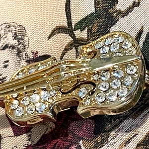 May include: A gold-toned violin-shaped brooch, encrusted with clear, sparkling rhinestones. The brooch has intricate detailing, including the violin's f-holes and strings. The background features a patterned fabric with a vintage aesthetic.