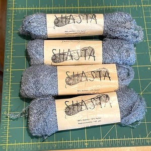 May include: Four skeins of blue mohair yarn, each wrapped in a tan paper label reading Shasta, 88% Mohair, 12% Nylon, and Approximately 145 yds. The yarn has a fuzzy, textured appearance and is arranged horizontally on a green cutting mat.